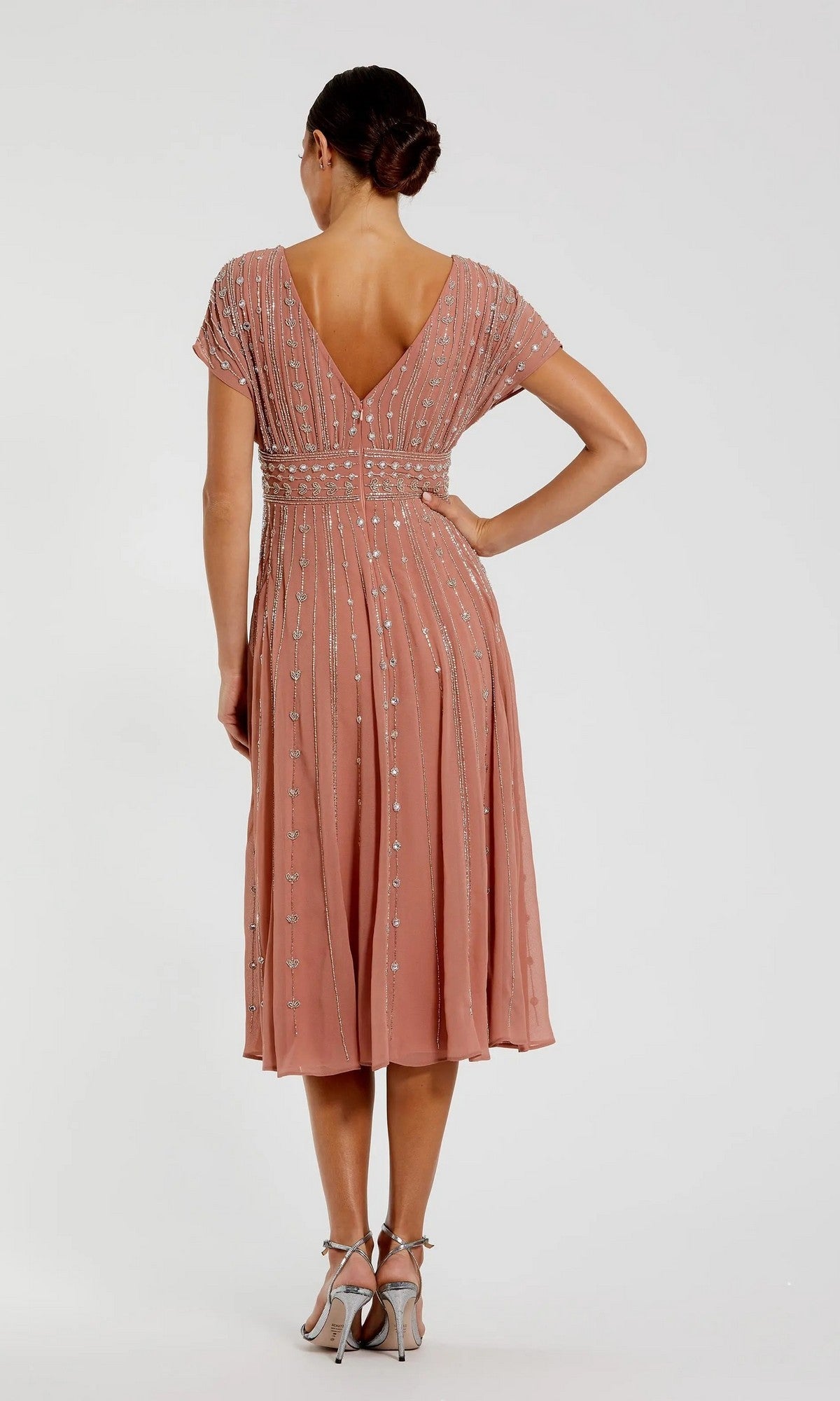 A woman faces away, dressed in the Midi Party Dress: Mac Duggal 94153—a peach, knee-length beaded midi dress with short sleeves and delicate vertical patterns—paired with silver heels, set against a plain light background.